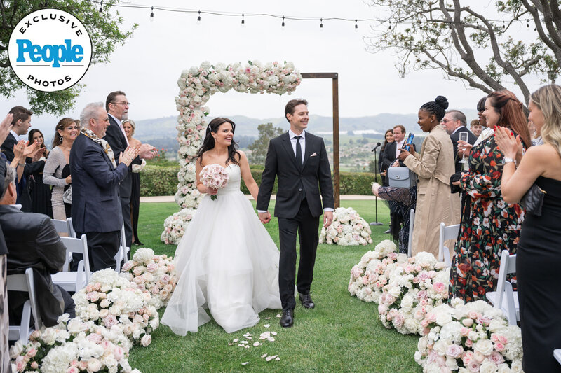 A newlywed couple walks hand in hand down the aisle at Spanish Hills, surrounded by cheering guests and stunning floral arrangements. The bride holds a bouquet of soft pink roses while the groom smiles proudly. This beautiful moment was captured by a wedding photographer in Los Angeles, showcasing love and celebration in a breathtaking outdoor setting.
