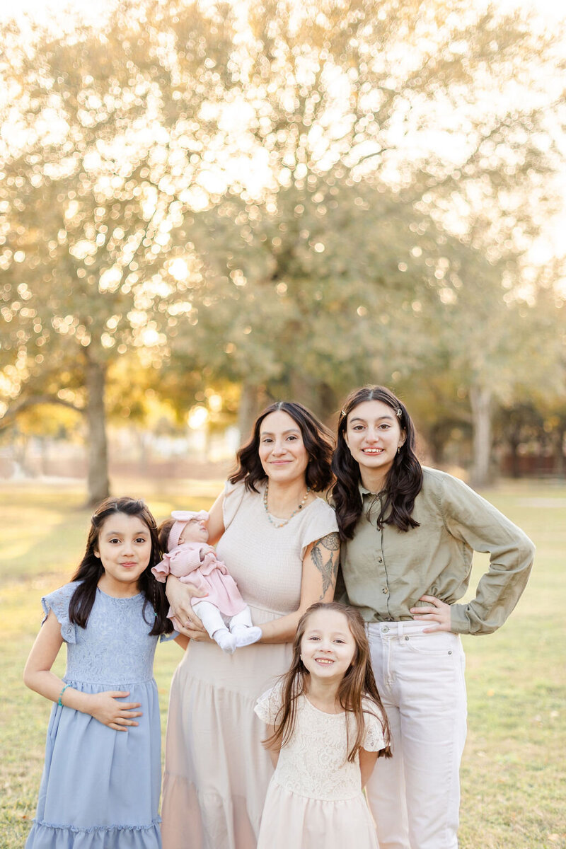 family portrait session in Pearland, Texas with the mother in cream, flowy dress while holding newborn in one arm and three daughters smiling at her side wearing a blue flowy dress, a pink flowy dress, and cream pants with an olive button down shirt