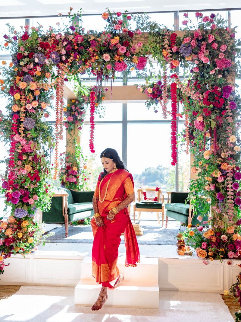 A person in a red and gold saree stands on steps under a floral arch adorned with vibrant flowers. The background shows a beautifully decorated room with seating and large windows.