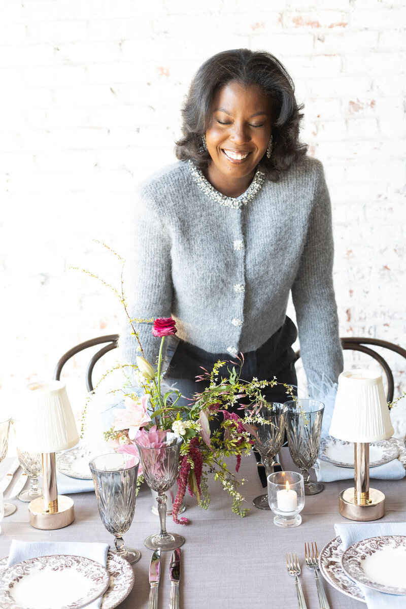 Photo of Tyler Albritton, a Georgia Planner in Atlanta, setting up a tablescape. She's smiling, and in front of her sits a pink floral centerpiece with greenery, small white candles, and mini lamps with white shades