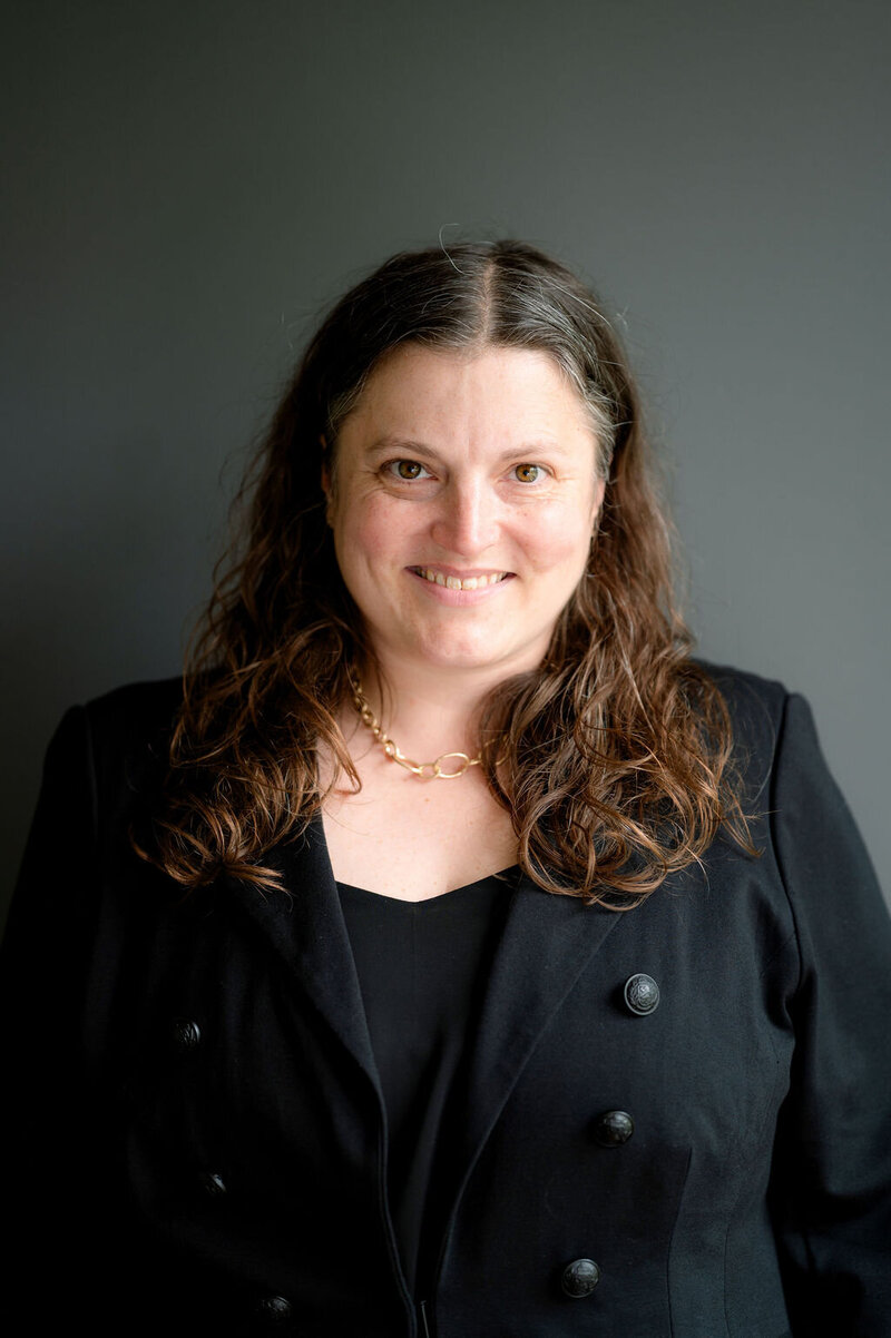 Jackie standing against gray background in black top and blazer