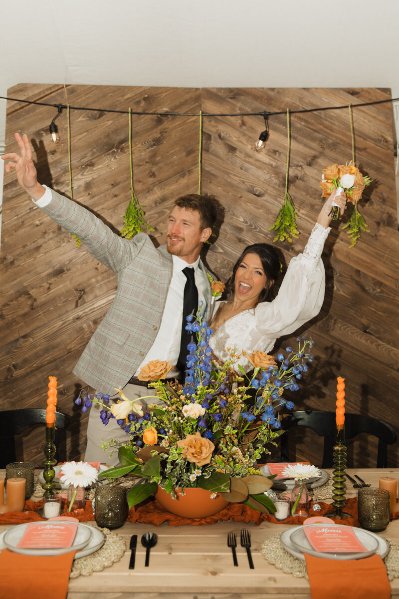 A bride and groom waving to their wedding guests  behind the wedding reception table.