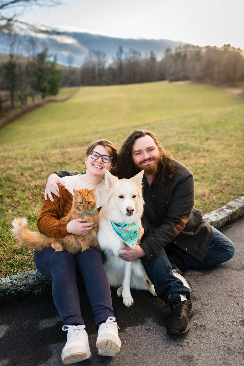 A Virginia elopement photographer sits on a curb leading to Cahas Mountain Overlook along the Blue Ridge Parkway. She sits next to her husband. An orange cat sits in her lap while their dog sits in between her and her husband.