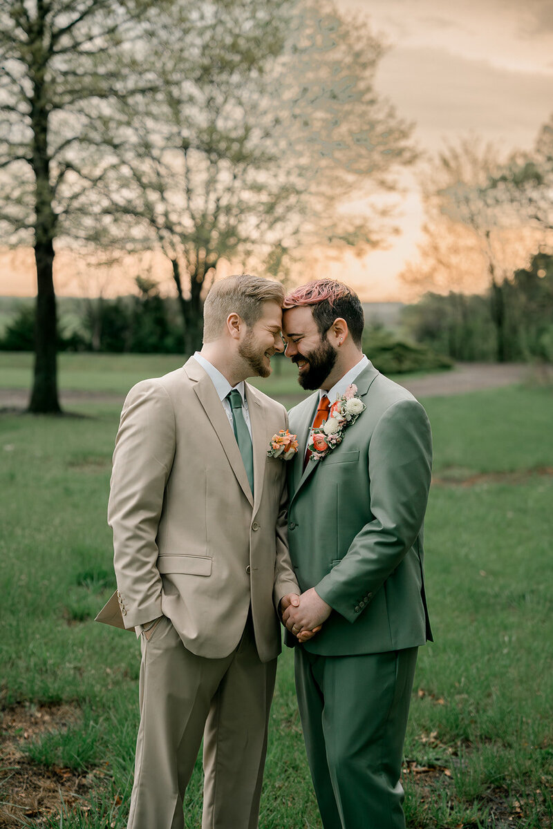 Grooms stand together holding hands and pressing their foreheads together outside during the sunset