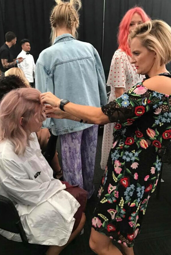 Hair artist Katy Reeve styling pastel pink hair backstage during a production, surrounded by models in vibrant outfits.