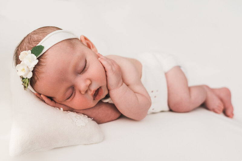 A peacefully sleeping baby lies on a soft white surface, wearing a white diaper and a white headband adorned with small flowers. The baby is resting on its side with a tiny pillow under its head.