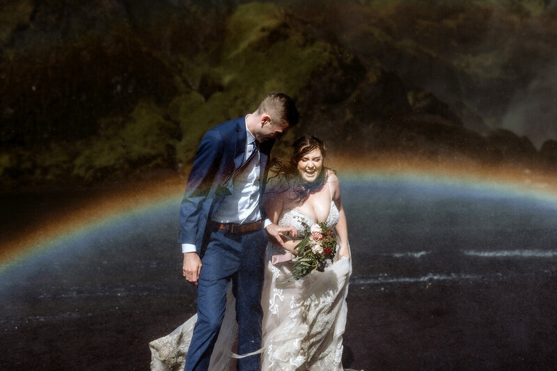Married couple is laughing while getting soaked under a waterfall surrounded by a rainbow in south Iceland on their wedding day
