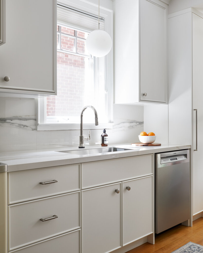 A bright airy kitchen with modern white cabinetry, wood flooring covered by a grey and white patterned rug, a stainless steel dishwasher, and a globe pendant lighting fixture hanging from the ceiling near a window