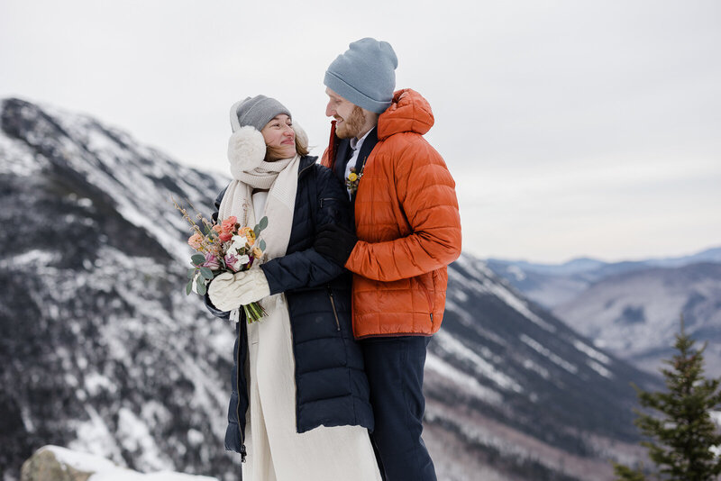 A couple smiles at one another during a snowy hiking elopement.