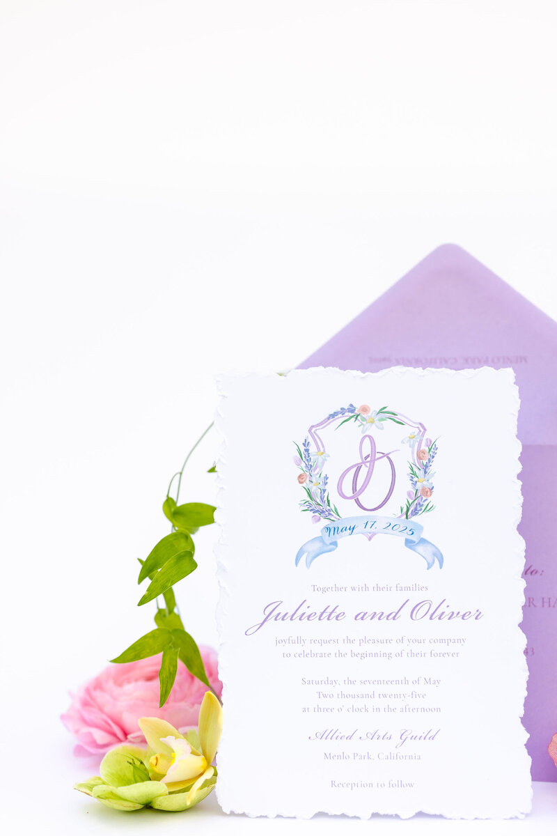 A delicate wedding invitation card with floral design details and calligraphy text, placed in front of a lavender envelope with pastel flowers, all set against a clean white background.