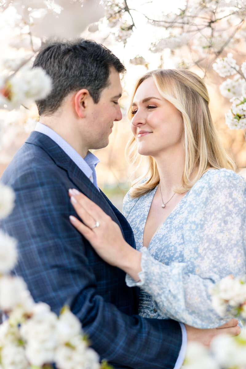 A couple stands closely together, smiling and surrounded by white flowering tree branches. The woman wears a light blue floral dress, while the man is in a dark suit. They gaze at each other affectionately.