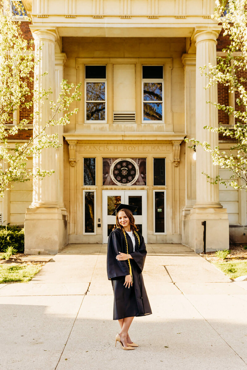 Beautiful brown haired woman crossing her ankles and touching her elbow while wearing her cap and gown for her graduation photo session in Green Bay