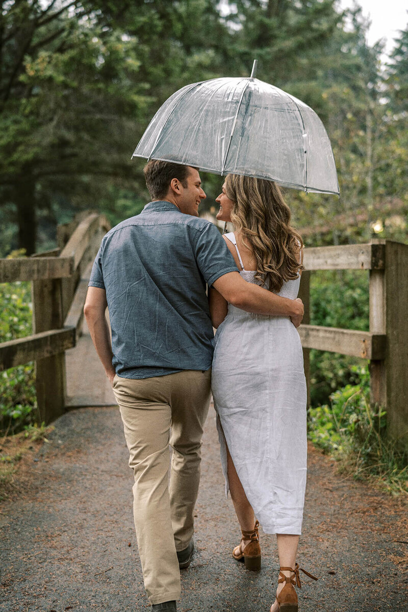 A couple share an umbrella and walk away looking at each other, representing the work of candid photographer Among the Oaks. 