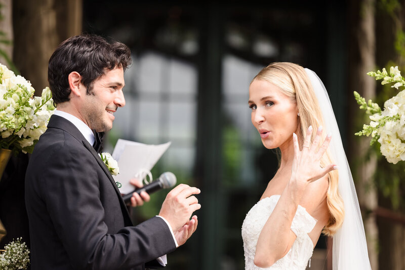 Bride playfully showing off wedding ring during ceremony as groom smiles with joy.