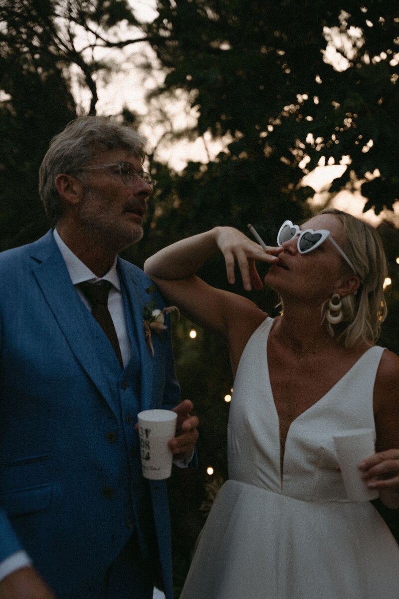 Mariés verre à la main et en train de fumer une cigarette en extérieur. Mariage à St Quay Portrieux en Bretagne par Kamescop.