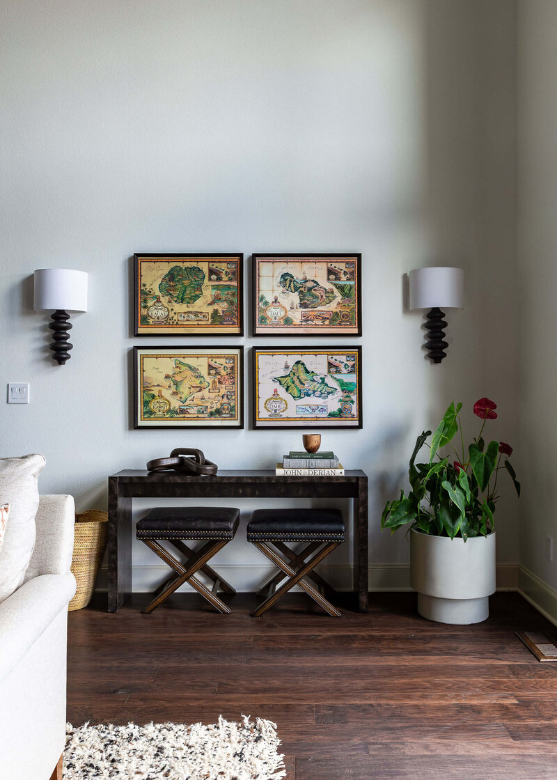 A table with books stacked on top, two stools beneath it and framed maps hanging on the wall above it.