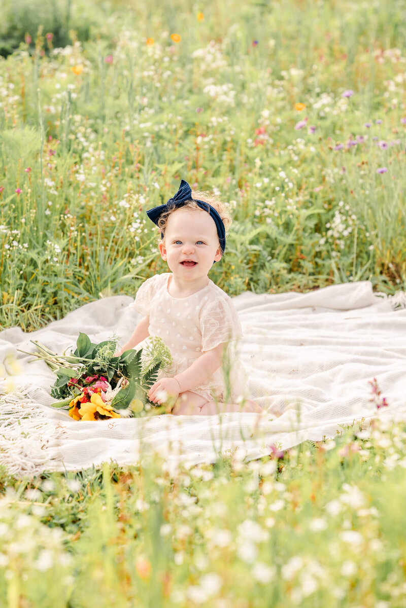A baby wearing a cream outfit and blue bow, sits on a white blanket in a flower field. She also holds on to a bouquet of flowers.