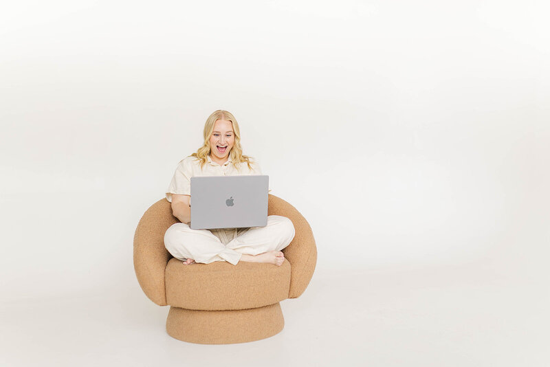 
Cheerful photographer sitting in a cozy chair, excitedly working on a laptop, showcasing a creative and engaging atmosphere in a bright, minimalistic setting.