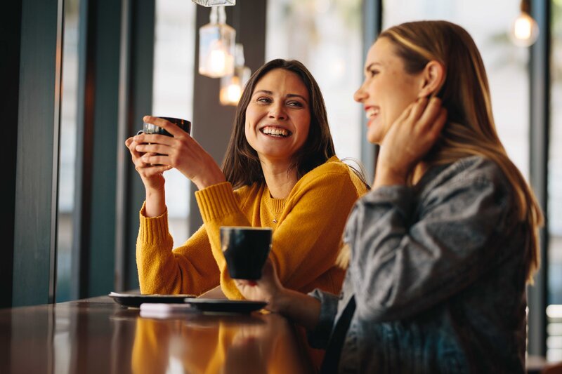 Two friends sit at a cafe countertop, each with a mug in front of them. They are smiling and laughing together.