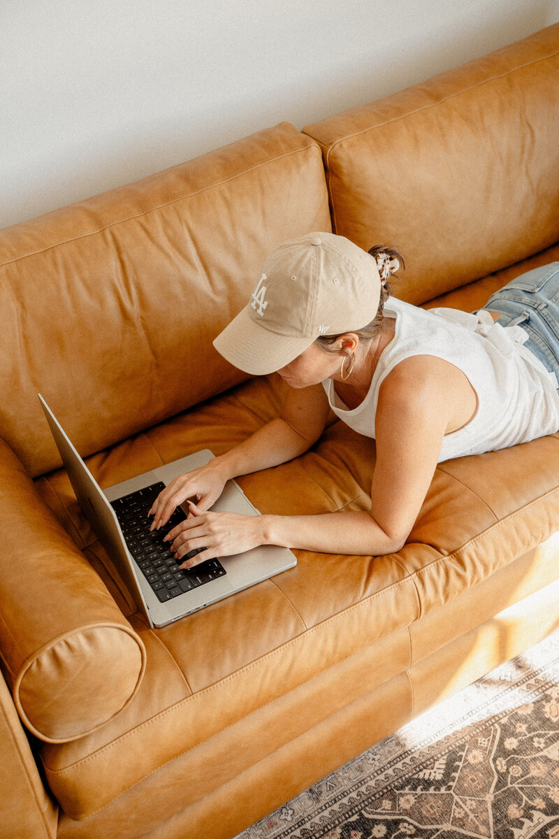 Woman wearing a ballcap laying on a leather couch typing on a laptop