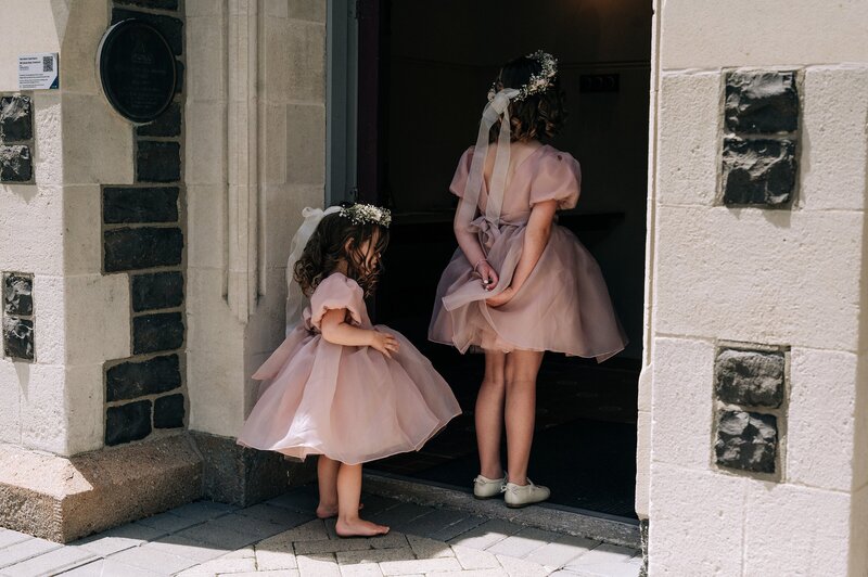 two flower girls in pink tulle dresses with flower crowns wait outside old church in christchurch