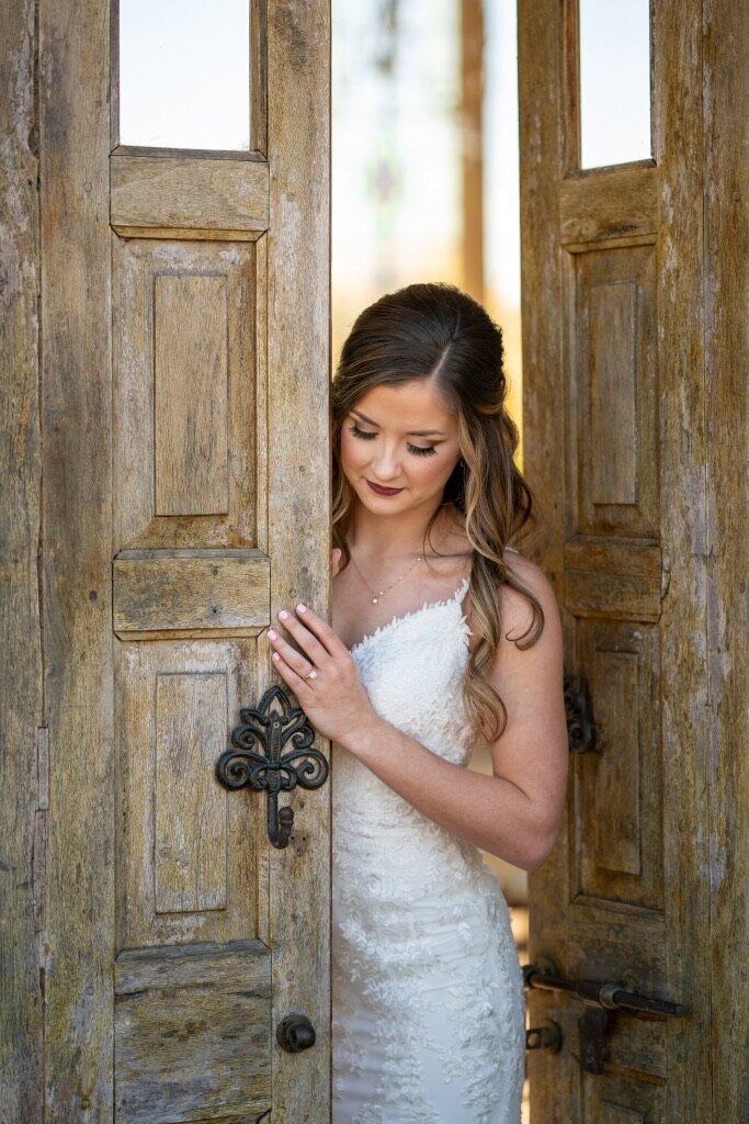The bride, with her best wedding hair accessories perfectly complementing her white dress, stands thoughtfully between rustic wooden doors.