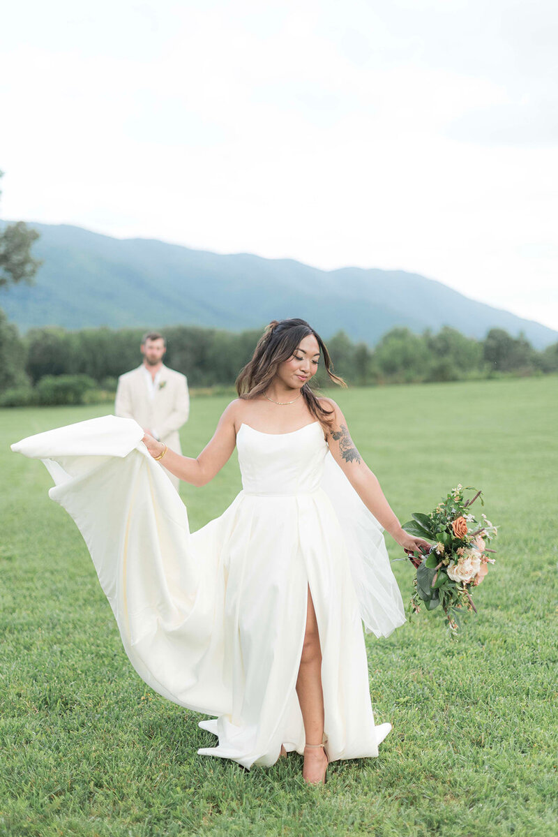 Beautiful bride poses with boquet while groom watches from a distance in front of dreamy scenic mountain background.