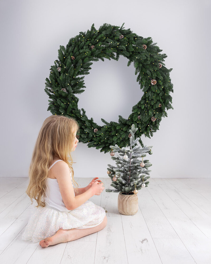 Image of a girl decorating a mini Christmas tree in front of a large Christmas wreath backdrop. Taken by Norwich Photographer Claire Howes. Christmas Minis Sessions Norwich.