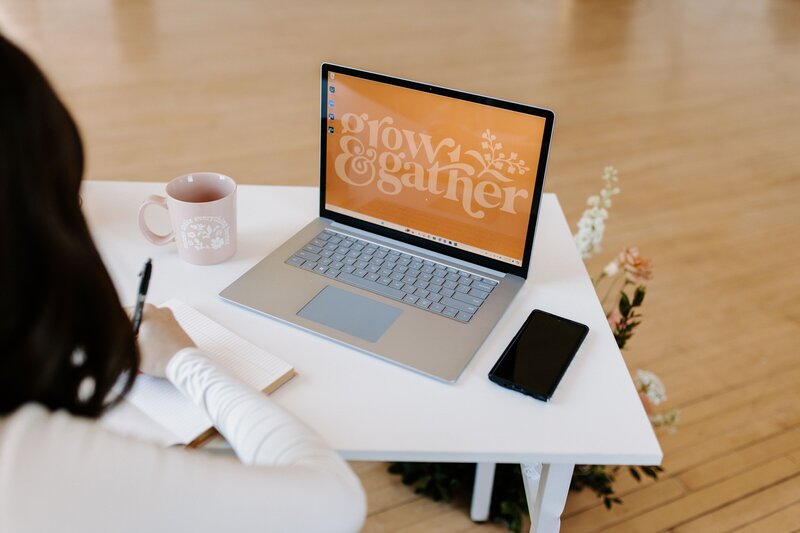 Woman with dark hair sitting at desk writting in notebook with laptop opena nd pink coffee mug