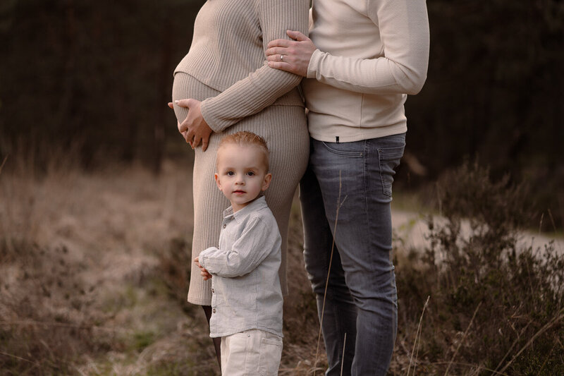 Grote broer staat voor zijn zwangere mama en papa en kijkt naar de lens.