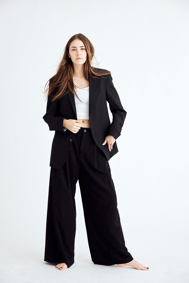 Stylish teen girl in an oversized black blazer and wide-leg trousers, paired with a white crop top, posing confidently in a minimalist white studio.