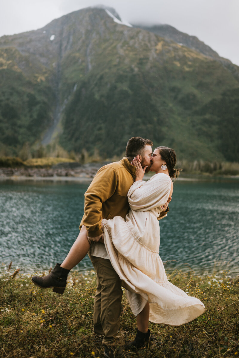 couple dip kissing in front of a mountain and lake with dramatic dress flowing in the wind during their engagement photos