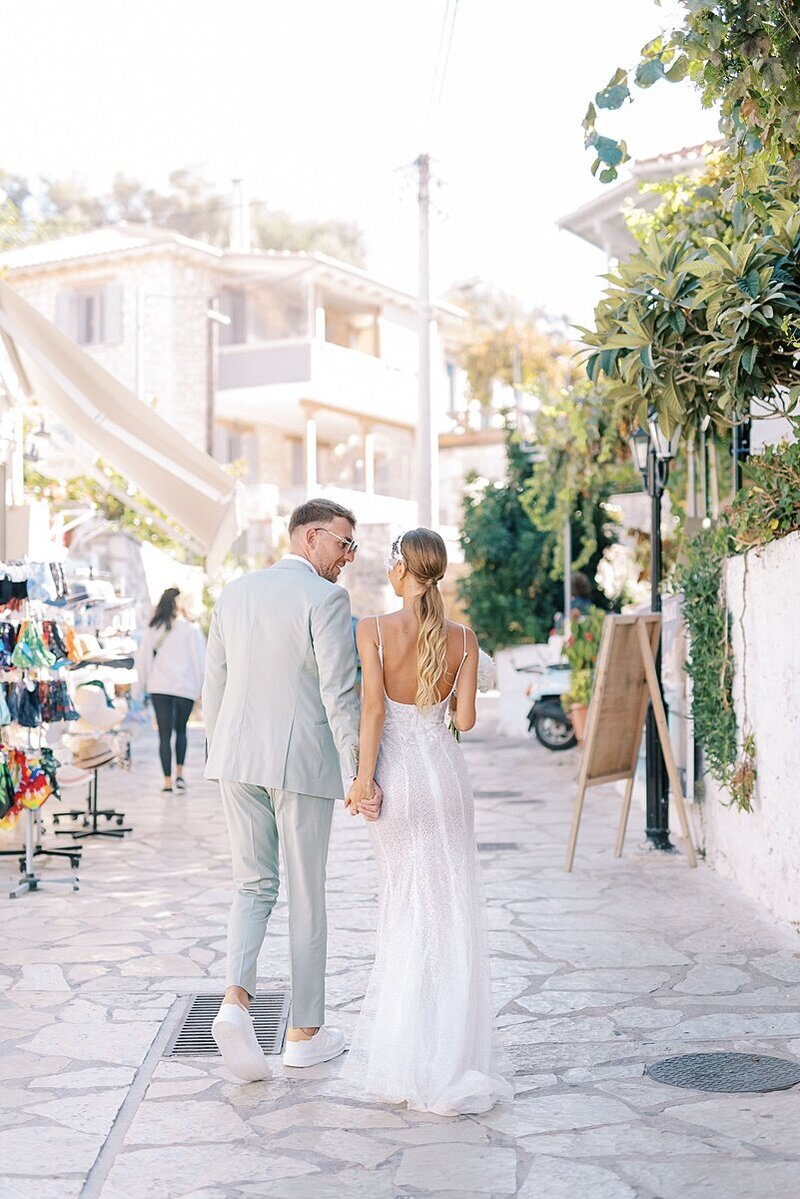 Bride and groom strolling hand in hand through a charming sunlit street in a coastal Greek village after their wedding ceremony