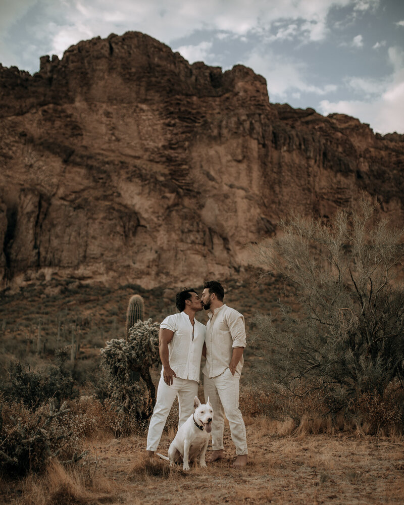 gay couple kissing in arizona desert with cliffs in white outfits with their dog