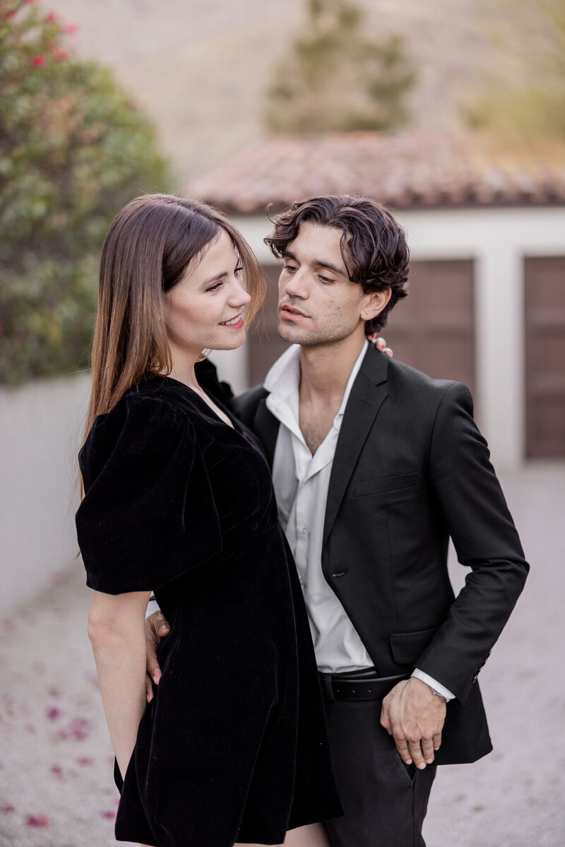 Stylish couple sharing a laugh on the sidewalk, dressed in modern black and neutral outfits with vintage city flair.
