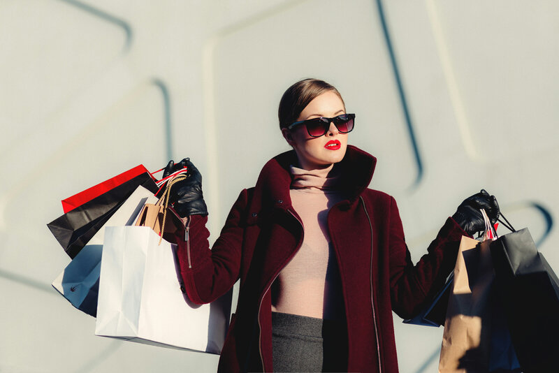 woman in a maroon jacket, black gloves, sunglasses, and bright red lipstick holding multiple shopping bags