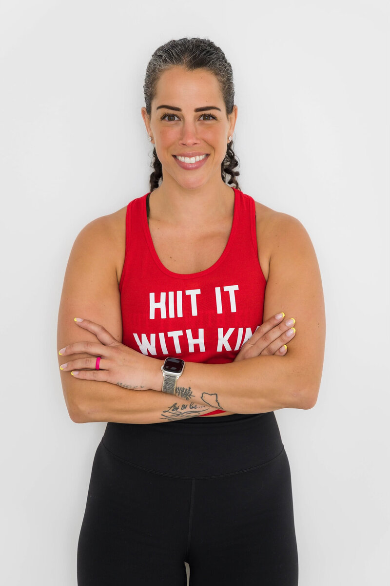 For weight loss, fitness, health & wellness. Kim is standing arms crossed in a Hiit It With Kim Red tank top with white letters smiling at the camera