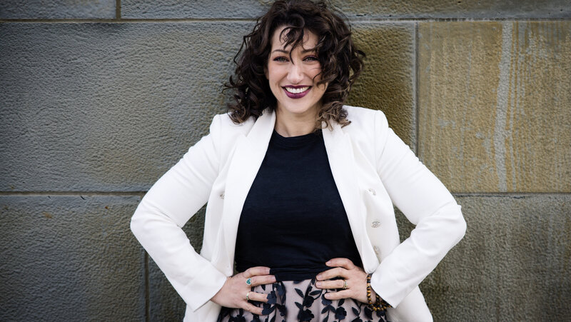 Woman in white blazer with hands on her hips smiling at the camera