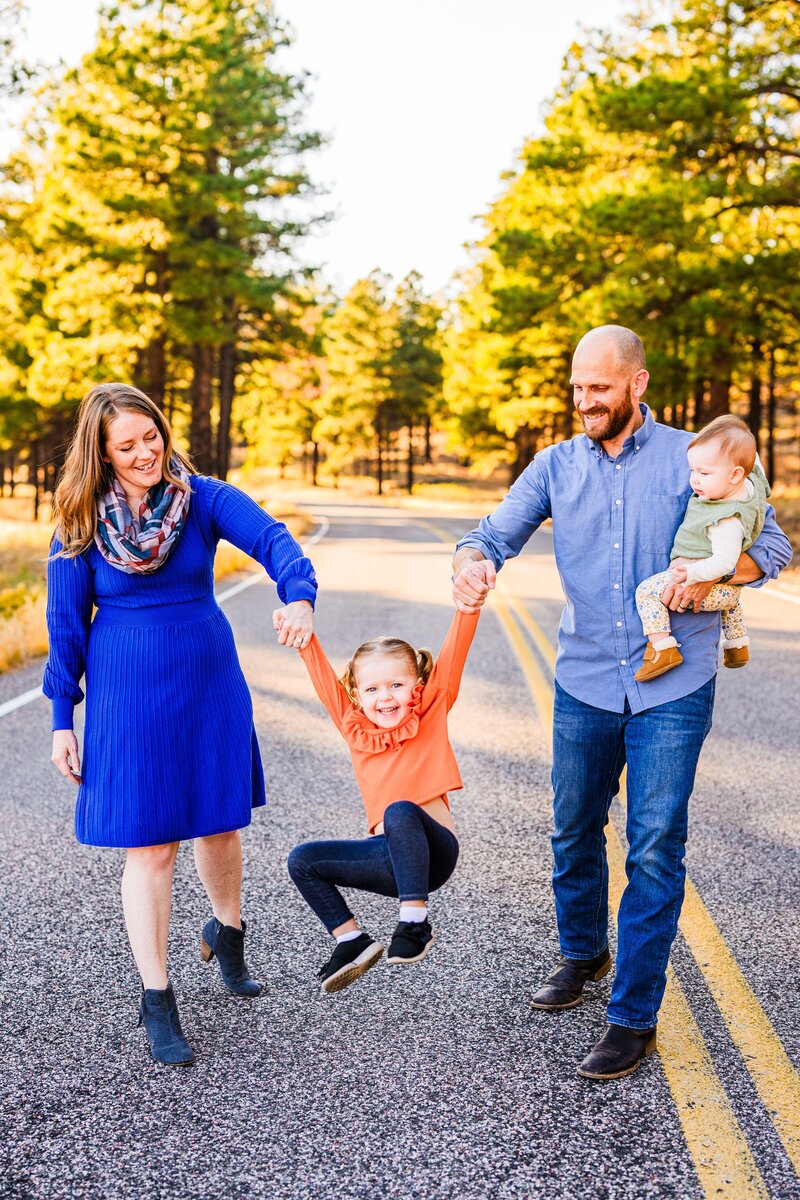 family with small children swinging girl on road with pine tree background golden hour in Flagstaff