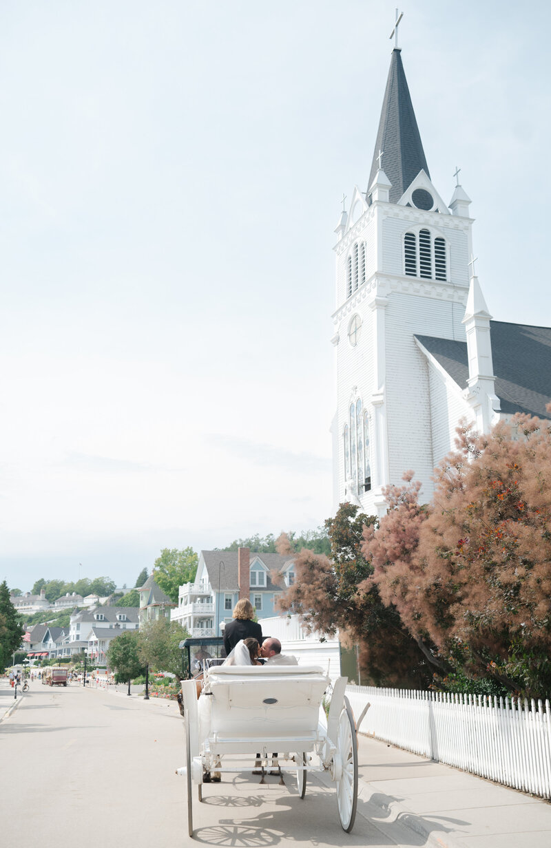 A bride and groom ride on a horse and carriage in front of a white Church at their wedding on Mackinac Island on a sunny summer day.