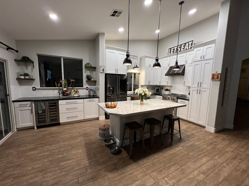 A newly renovated kitchen with faux wood tiled floors, a large island in the center with two black pendant lights and black barstools. The cabinets are white with black hardware and the appliances are stainless steel.