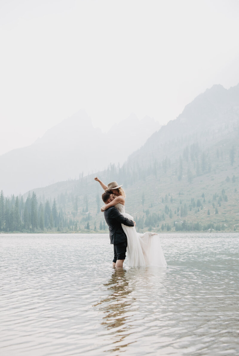 string lake wedding with bride and groom in the water and the groom lifting up his bride as she raises her arm in the air captured by jackson hole photographers with the grand tetons in the distance
