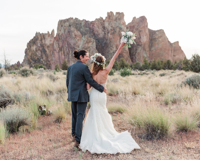 Couple just married in wedding attire facing away from the camera towards mountains in the distance. The bride is wearing a bohemian flower crown with pastel assorted flowers and is holding her bouquet in the hair triumphantly.