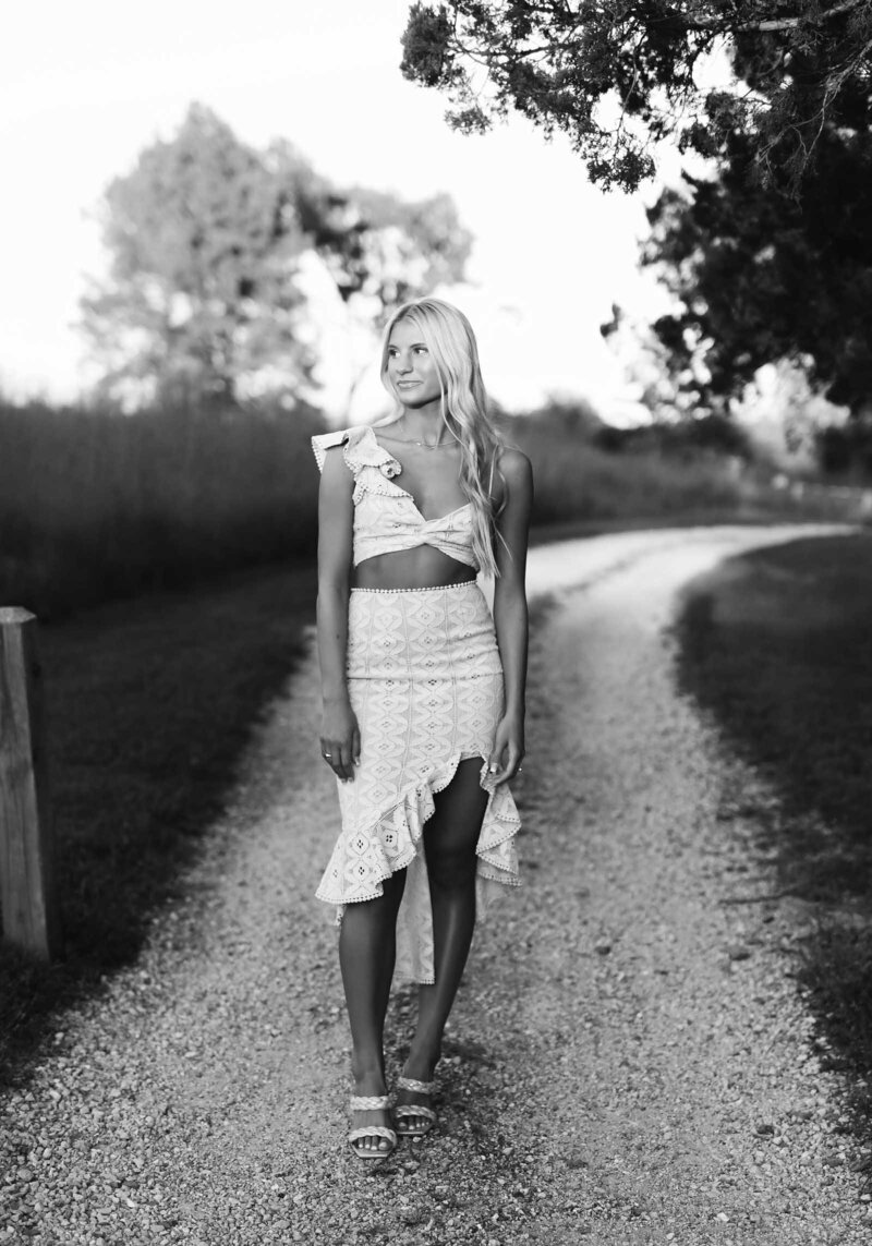 A woman with long, wavy hair stands outdoors in a grassy area, captured beautifully by a St. Louis photographer. She is wearing a light-colored, sleeveless, two-piece outfit with ruffled shoulder details and an asymmetrical skirt. Looking down and smiling, the black-and-white image exudes timeless elegance.