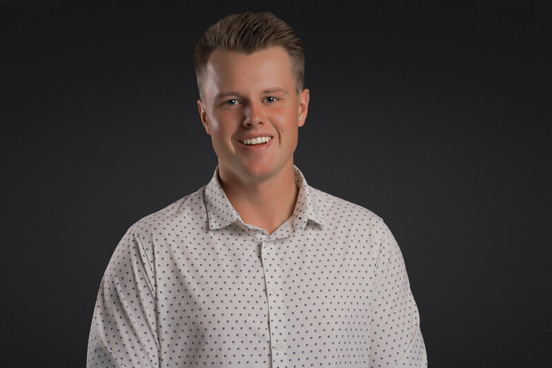 A young man with short, neatly styled hair smiles confidently at the camera while wearing a light-colored, patterned button-down shirt.