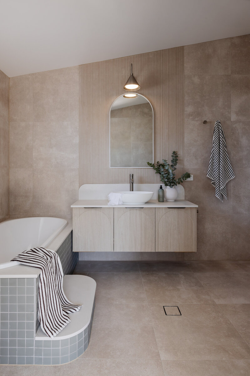 Minimalist bathroom with neutral tones, crafted by a Newcastle interior designer, featuring a bathtub, striped towels, and a modern vanity with a arched mirror.