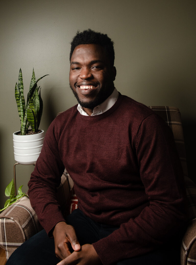 A Black man in a sweater smiles for a photo next to plants.