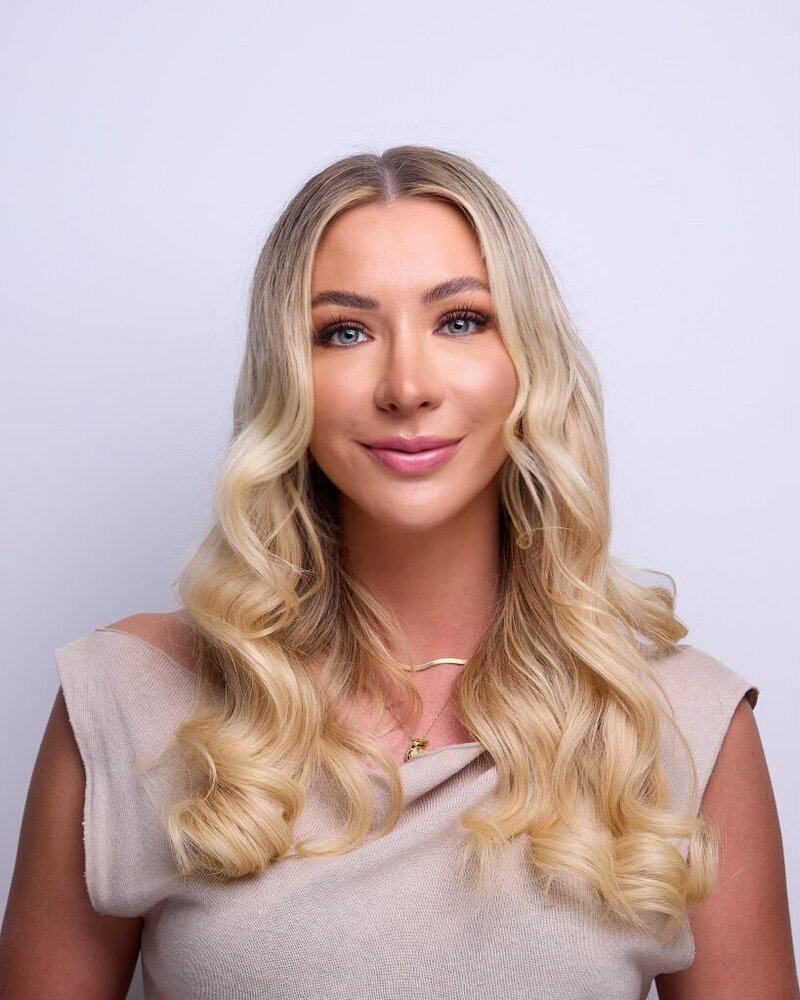 Portrait of a woman with blonde hair. She is smiling and wearing clothing. The photo focuses on her face, neck, and shoulders.