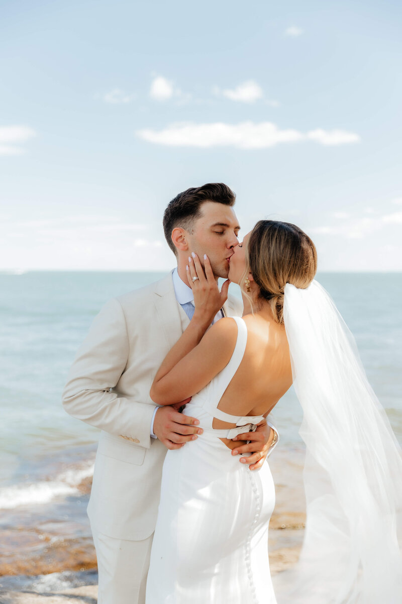 modern couple shares romantic kiss by the ocean, bride in sleek backless dress and veil, candid wedding day moment