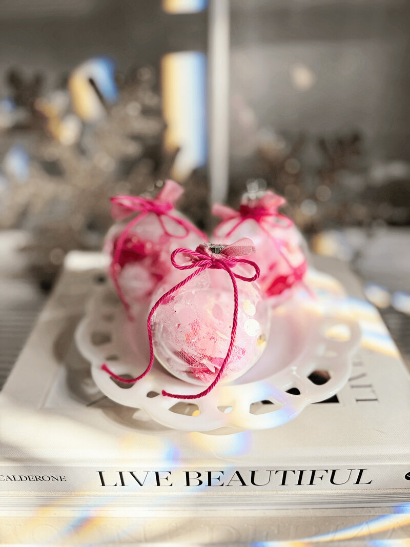 Three clear holiday ornaments filled with pink material and tied with pink ribbons sit on a white decorative plate, placed on a book titled "Live Beautiful," showcasing Sweet Pea Ink Creative’s charming style.